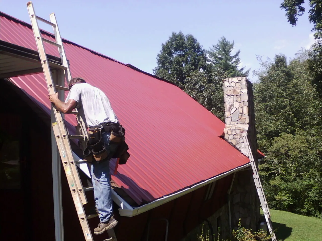 Roofer on a ladder beside a red metal roof and stone chimney — Soffit & Fascia Repair by Roofing Brothers in Tarzan